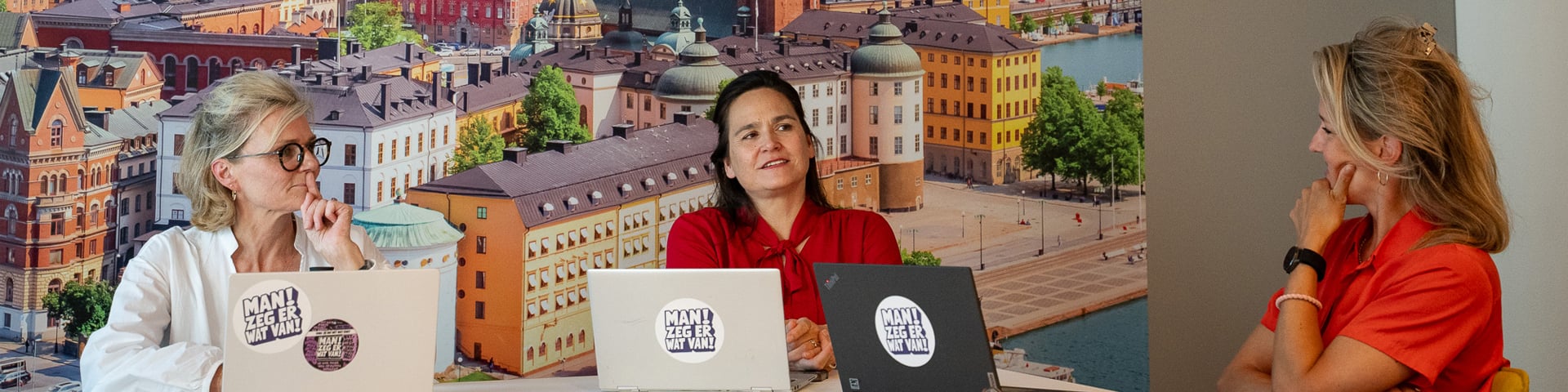 drie vrouwen zitten met laptops aan een tafel en kijken elkaar aan, met op de achtergrond een foto van Den Haag