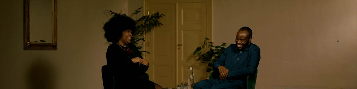 Lucelle Comvalius en George Adegite zitten naast elkaar in een donkere ruimte. Ze kijken elkaar lachend aan.