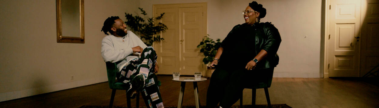 Safi Graauw en Shirma Rouse zitten naast elkaar op stoelen in een grote kamer. Ze kijken elkaar lachend aan.