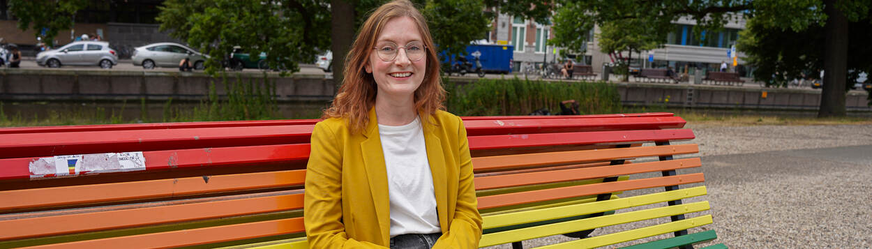 Lieke Schrijvers is een jonge witte vrouw met rood haar en een ronde bril. Ze draagt een geel jasje met daaronder een wit shirt en een donkergrijze broek. Ze zit op een regenboogbankje in een park en kijkt lachend in de camera.