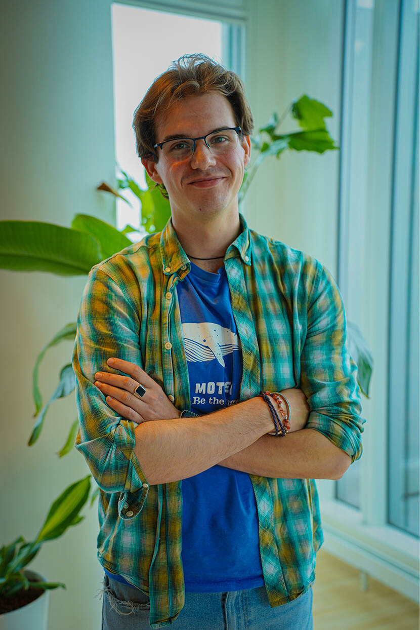 Bas van den Berg is een witte man met kort bruin haar en een bril. Hij staat met zijn armen over elkaar geslagen voor een plant en een raam en kijkt met een glimlach in de camera. Hij draagt een bloes met blauw-geel-witte ruiten en daaronder een blauw shirt.