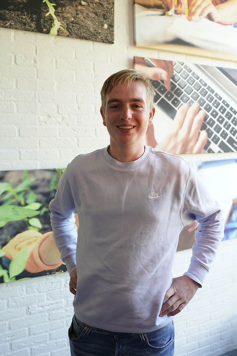 Leonard, een blonde jongen in een lichtblauwe trui en spijkerbroek, poseert voor een muur met foto's in zijn school - Accent Praktijkonderwijs in Nijkerk. Hij heeft zijn handen in zijn zij en kijkt lachend in de camera.