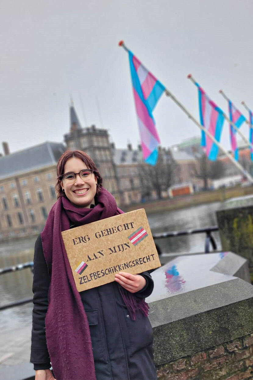Amber draagt een bril en een donkerpaarse sjaal. Die poseert voor de Hofvijver bij het Binnenhof in Den haag. In diens handen heet Amber een bord met de tekst 'Erg gehecht aan mijn zelfbeschikkingsrecht'. Op de achtergrond aan de rand van de vijver is een rij transvlaggen te zien (blauw, roze, wit, roze, blauw).