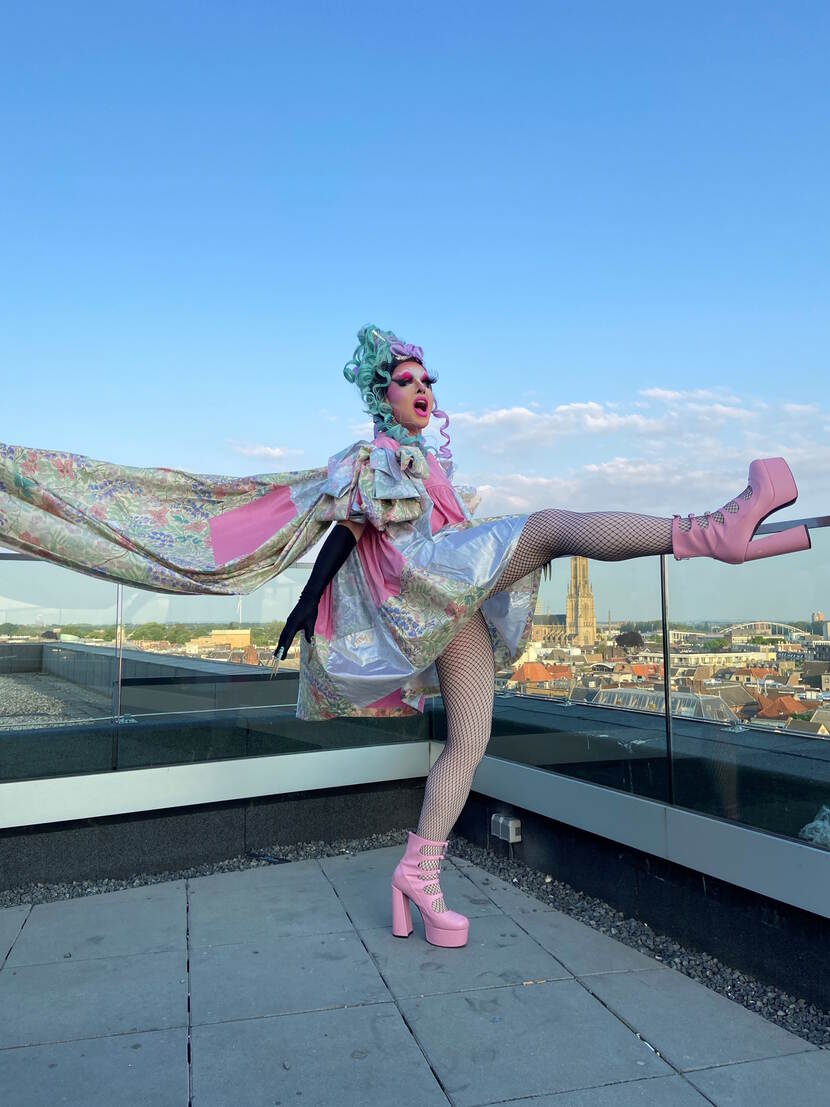 Nikki Pleasure is een drag queen en staat uitbundig gekleed op een dak met haar rechter been omhoog. Ze draagt grote roze hakken.