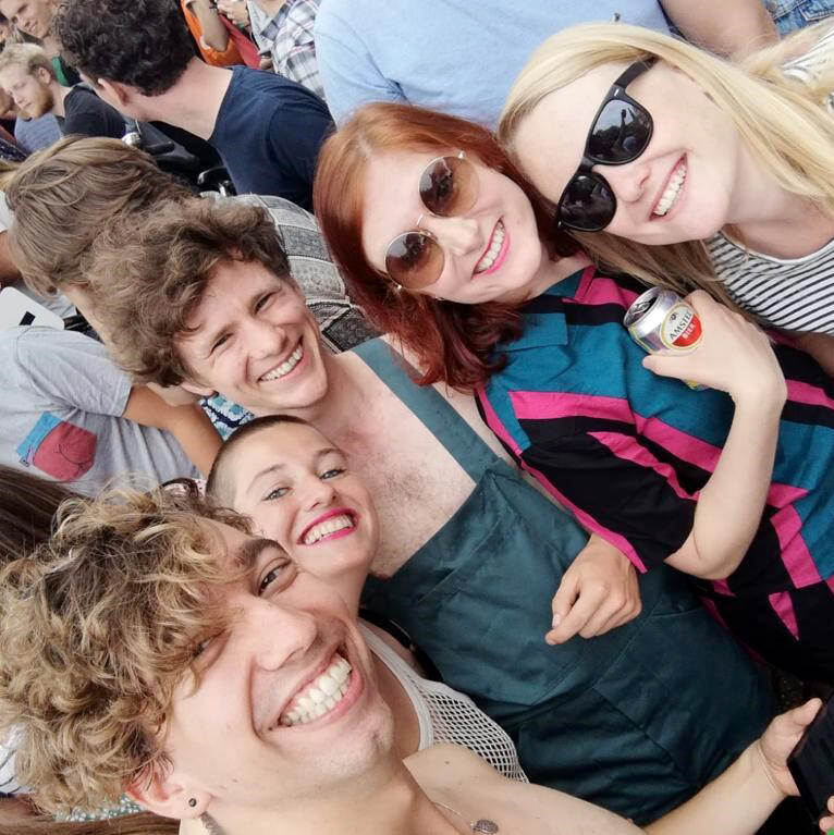 Lieke Schrijvers met haar vrienden op de pride in Amsterdam. De foto is een selfie en toont van links naar rechts een man met bruin haar, een vrouw met geschoren donker haar en roze lippenstift, een man met bruin haar en een donkergroene tuinbroek, een vrouw met rood haar en een zonnebril en een vrouw met lichtblond haar en een zonnebril. Alle personen op de foto kijken lachend in de camera.