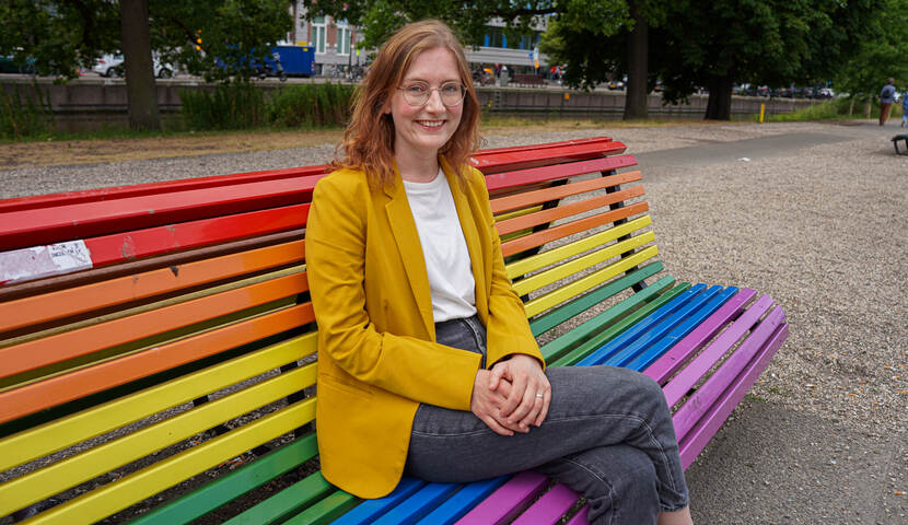 Lieke Schrijvers is een jonge witte vrouw met rood haar en een ronde bril. Ze draagt een geel jasje met daaronder een wit shirt en een donkergrijze broek. Ze zit op een regenboogbankje in een park en kijkt lachend in de camera.